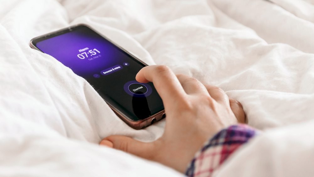 A hand reaching for a phone alarm on crumpled white bed sheets
