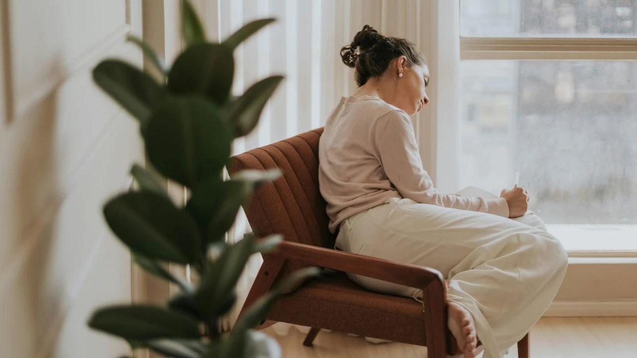 A woman sitting alone in an armchair by a large window at home, writing in a notebook