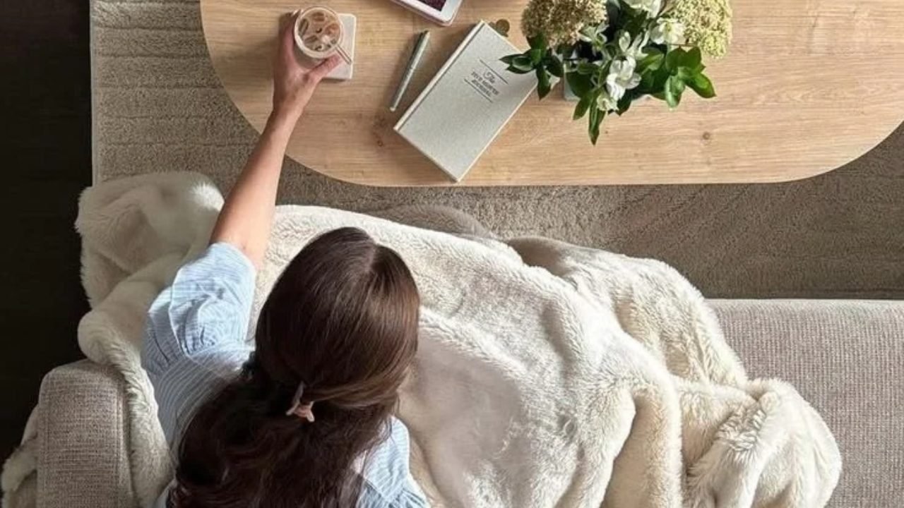 A woman with long dark hair resting on a sofa under a cream blanket, reaching for a drink on the coffee table beside a journal and white flowers