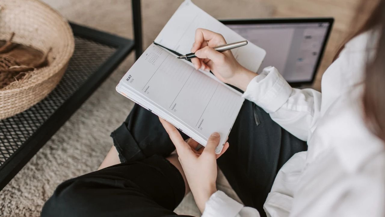 A woman in a white shirt writing in a weekly planner on her lap, with a laptop open beside her