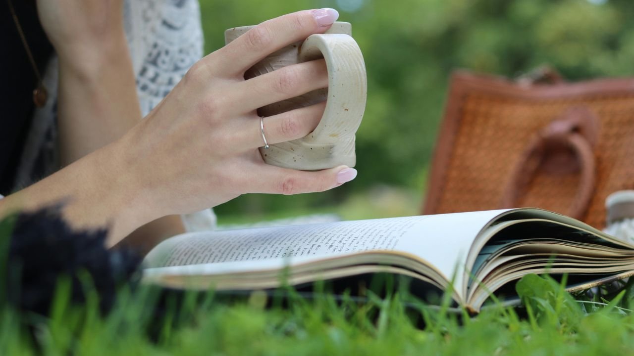 A hand holding a ceramic mug beside an open book on the grass